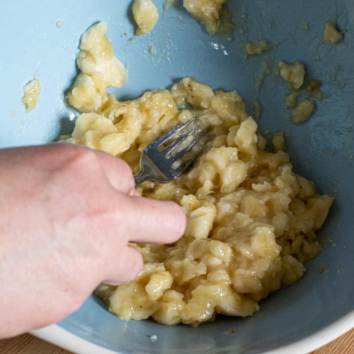 womans hand holding a fork and mashing bananas in a blue mixing bowl