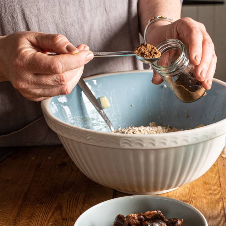 woman in grey spooning spices from a small glass jar into a large blue and white mixing bowl