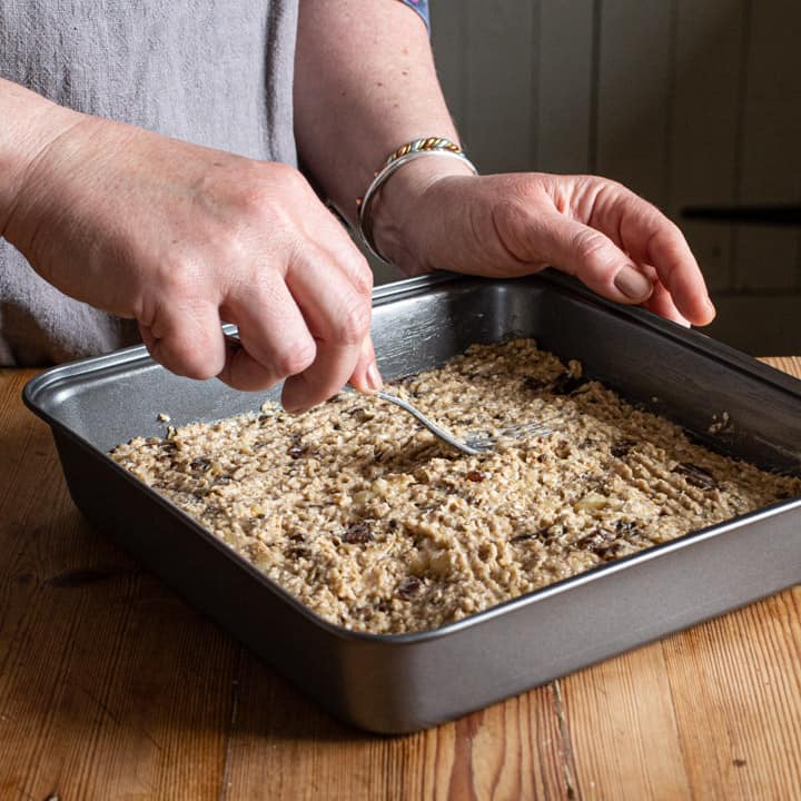 woman in grey smoothing down flapjack mixture into a square baking tin