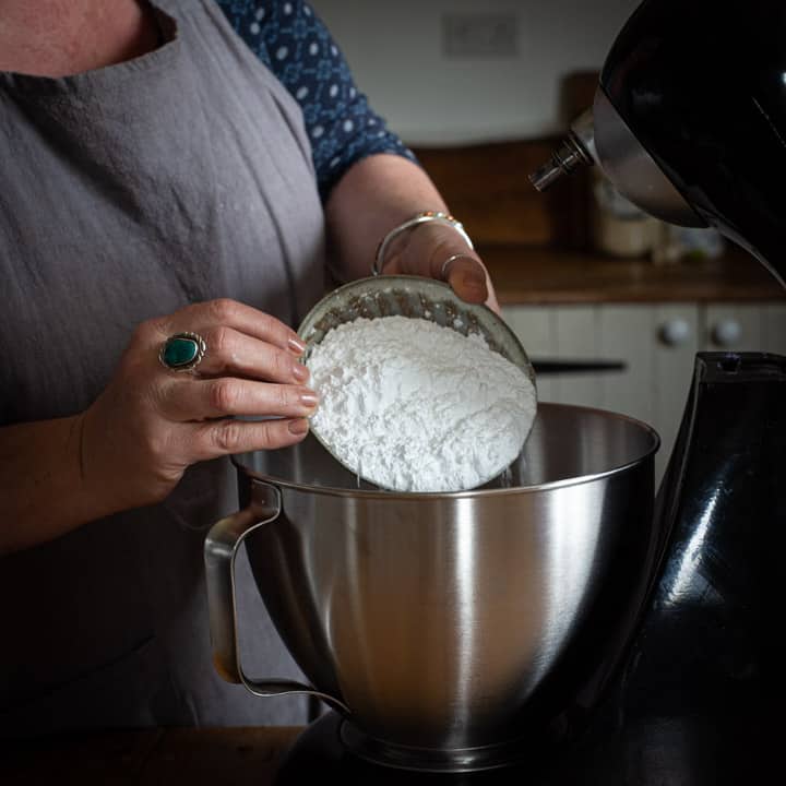 woman in grey tipping a bowl of icing sugar into a large mixing bowl