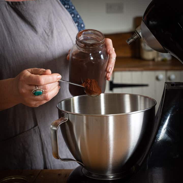 woman in grey spooning cocoa powder form a glass jar into a silver mixing bowl