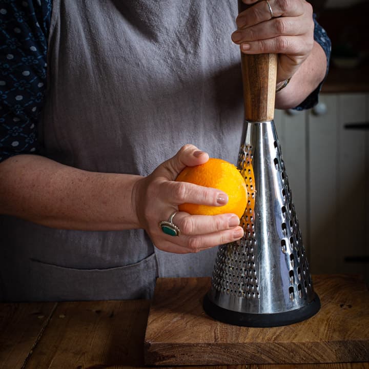 woman in grey grating an orange on a tall wood and metal grater