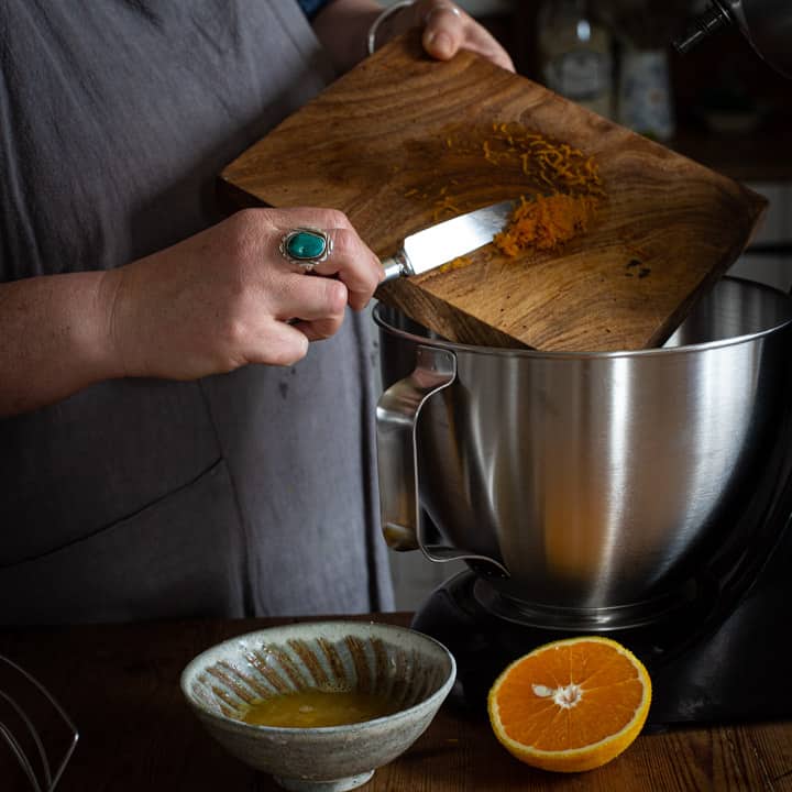 womans hands scraping frsh orange zest from a wooden board into a silver mixing bowl