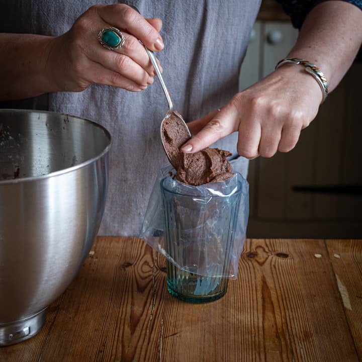 womans hands scraping chocolate orange frosting from a silver spoon into a glass lined with a piping bag