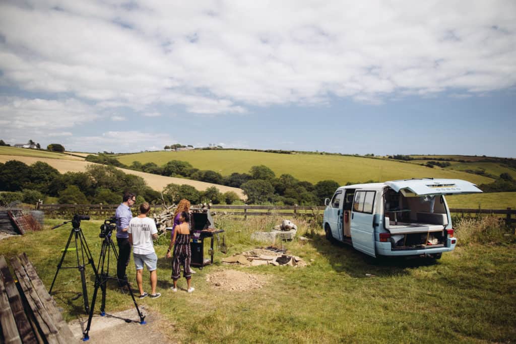 Landscape with fields. A campervan is sat in the field at the front together with a group of four people and camera equipment.  