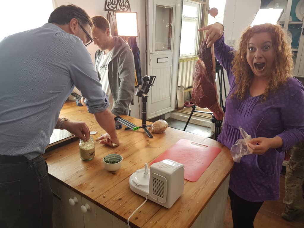 Hedgecombers Kitchen presenter making a surprised face holding a huge uncooked steak stood in kitchen with film crew and lighting 