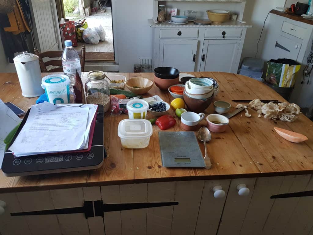 Cluttered kitchen table with ingredients, bowls, weighing scales, paperwork on the wooden surface