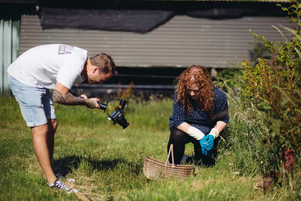 Presenter of Hedgecombers Kitchen crouching beside basket in allotment being filmed by man with camera