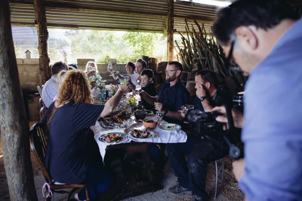 Group of people sat around table full of food raising their glasses while being filmed