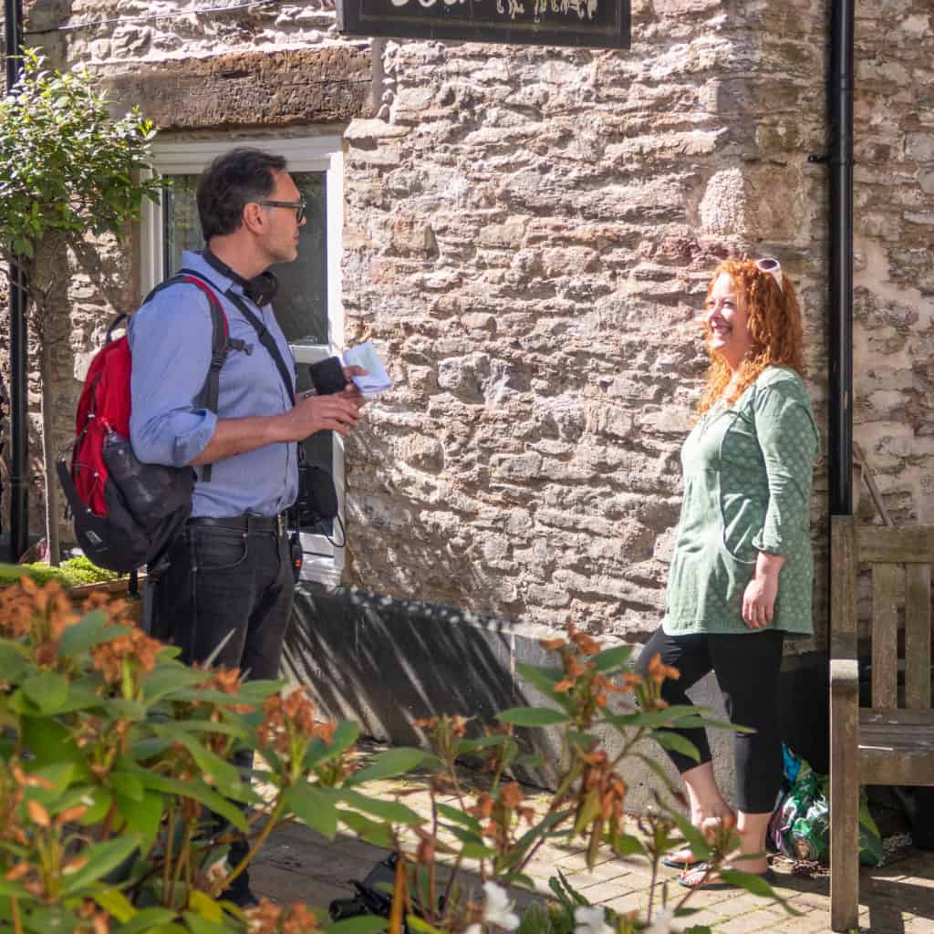 Presenter stood beside stone cottage smiling at man with headphones around his neck and rucksack