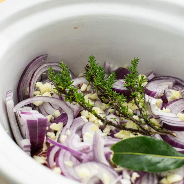 inside shot of a white slow cooker with chopped red onions, chopped garlic and fresh thyme and a bay leaf