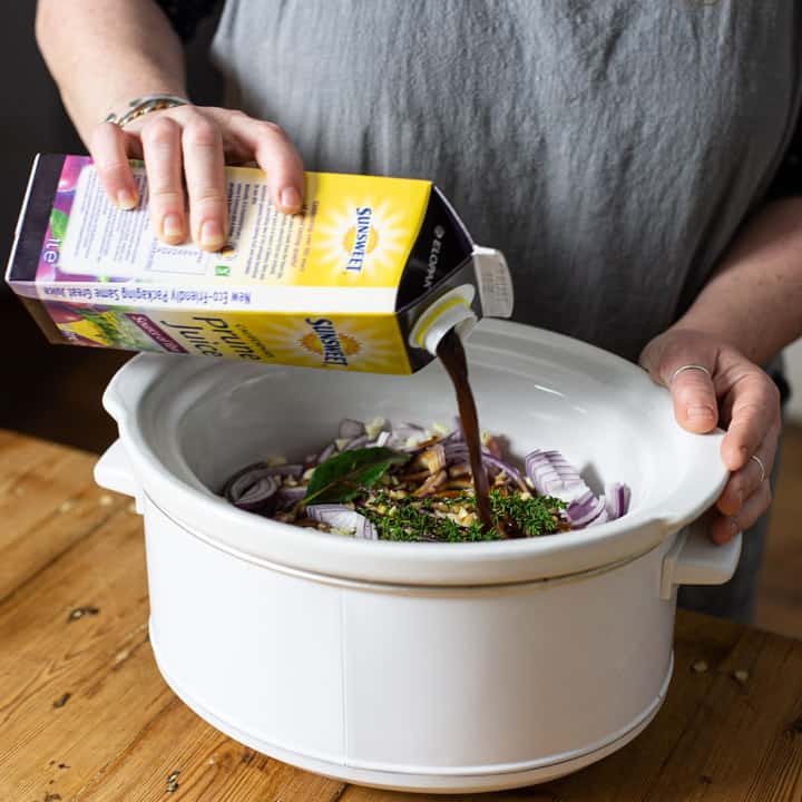 woman in grey jumper pouring prune juice from carton into a white slow cooker bowl with rabbit