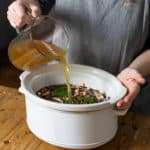 woman in grey jumper pouring hot stock into a white slow cooker bowl with rabbit
