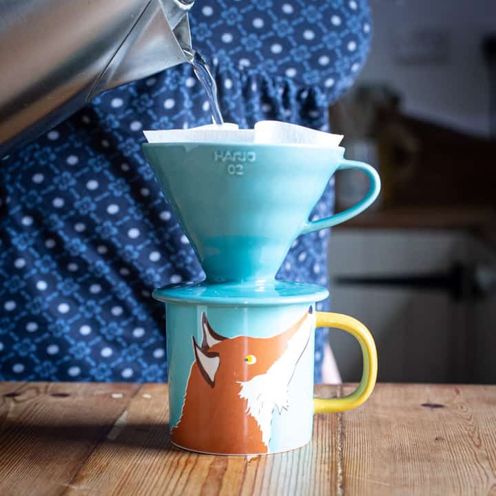 woman pouring water from a kettle into turquoise coffee maker over a fox cup