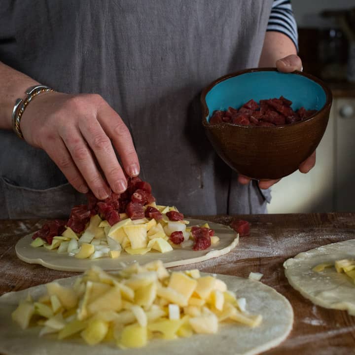 three discs of pastry on a wooden surface, piled up with raw swede, potato, onion and steak ready to be crimped into pasty's