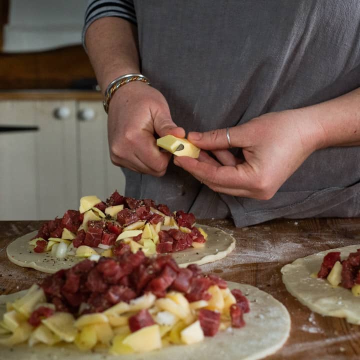 womans hands chopping a piece of butter up over the top of three pasties that are about to be crimped