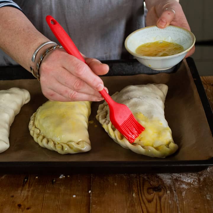 womans hands brushing beaten eggs with a red silicone brush onto three uncooked Cornish pastys on a black baking tray