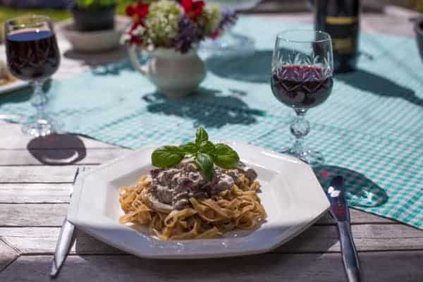 sunny wooden table with glasses of red wine, flowers and a plate of vegetarian mushroom pasta 