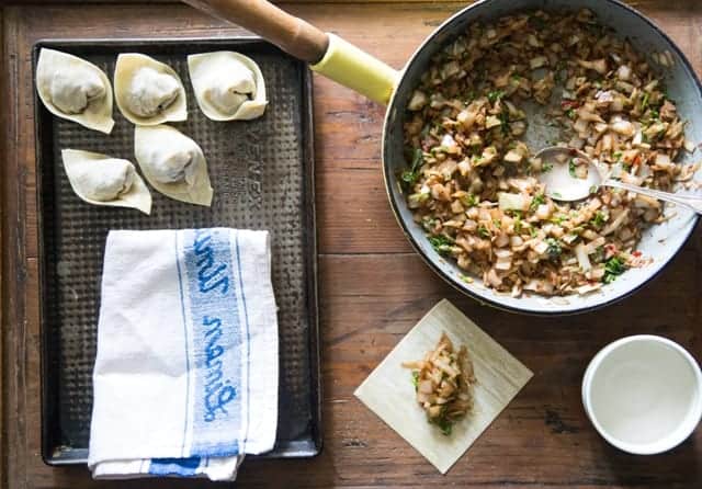 wooden table with a metal tray, tea towel and frying pan with vegetarian wontons being made at every stage