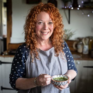 woman with red hair and grey apron holding a small bowl of bright green wild garlic soup in a kitchen