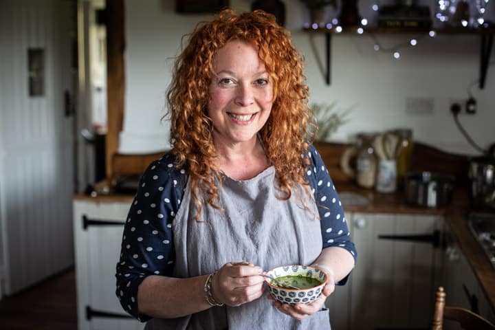 woman with red hair and grey apron holding a small bowl of bright green wild garlic soup in a kitchen