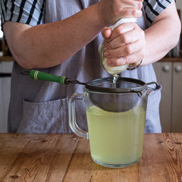 womans hands squeezing elderflower syrup into a old fashioned sieve over a glass jug