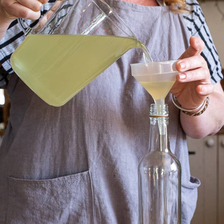 woman in grey pouring elderflower syrup from a glass jug into a a small funnel and glass bottle