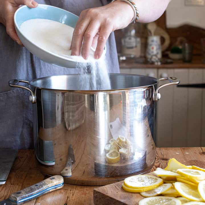 woman pouring sugar from a pale blue bowl into a large saucepan with slices of lemon around the base