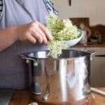 womans hands placing elderflower blooms into a large silver saucepan from a small white bowl