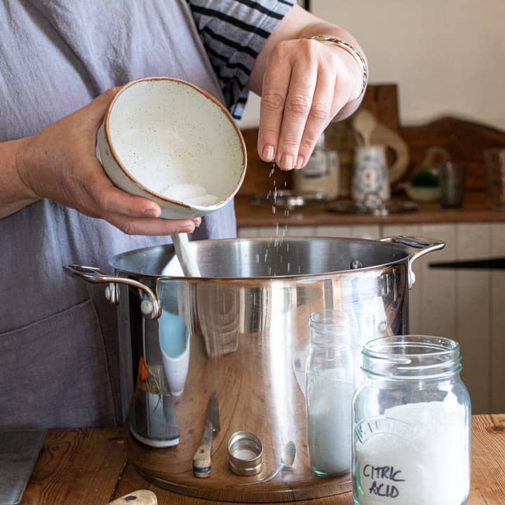 woman sprinkling citric acid from a small white bowl into a large silver saucepan