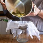 womans hands pouring elderflower syrup from a large silver saucepan into a muslin lined sieve