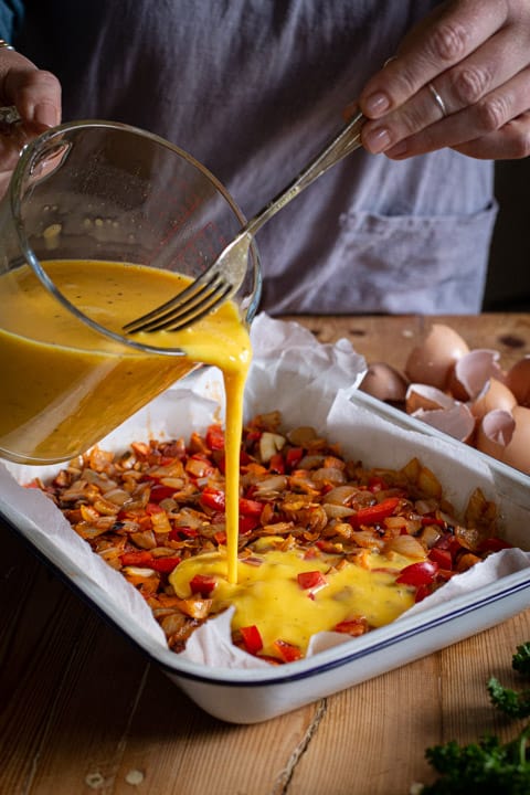 glass jug of beated eggs being poured into a white baking tin filled with frittata ingredient