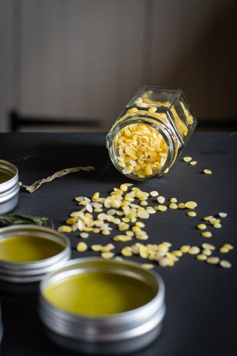 black background with a small glass jar on its side spilling small beeswax beads and 2 small tins of plantain salve