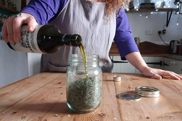 woman pouring olive oil into a glass jar half filled with plantain leaves in a rustic kitchen