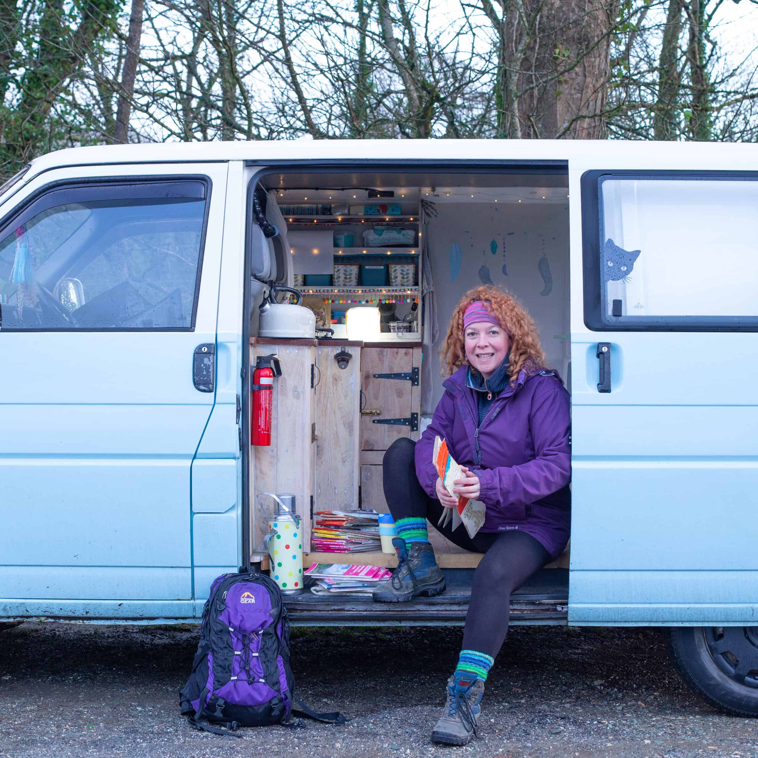 woman with red curly hair sat in pretty blue and white campervan with hiking gear, backpack and paper maps