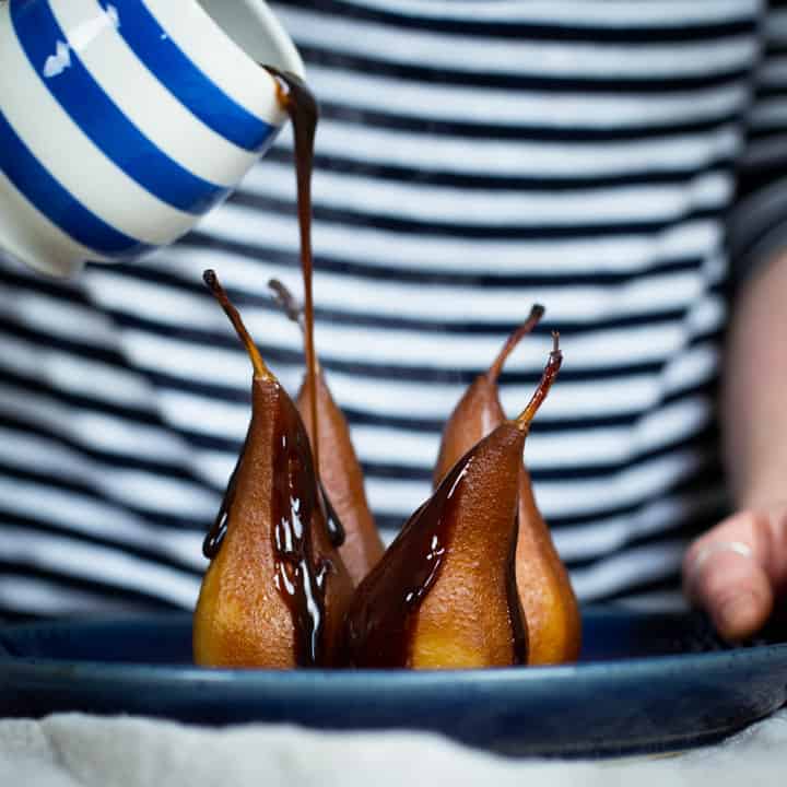 woman in blue and white stripy top pouring sweet prune sauce from a blue and white stripy jug over 4 poached pears on a blue plate