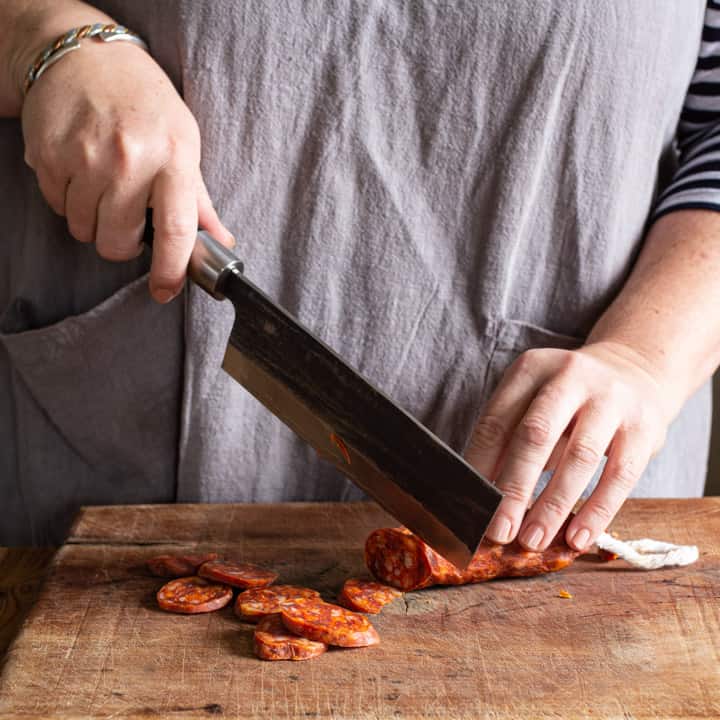 womans hands chopping chorizo on wooden chopping board with large Japanese knife