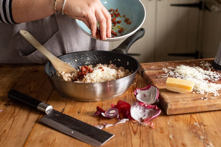 a messy kitchen risotto scene with womans hands tipping diced chorizo into the risotto pan