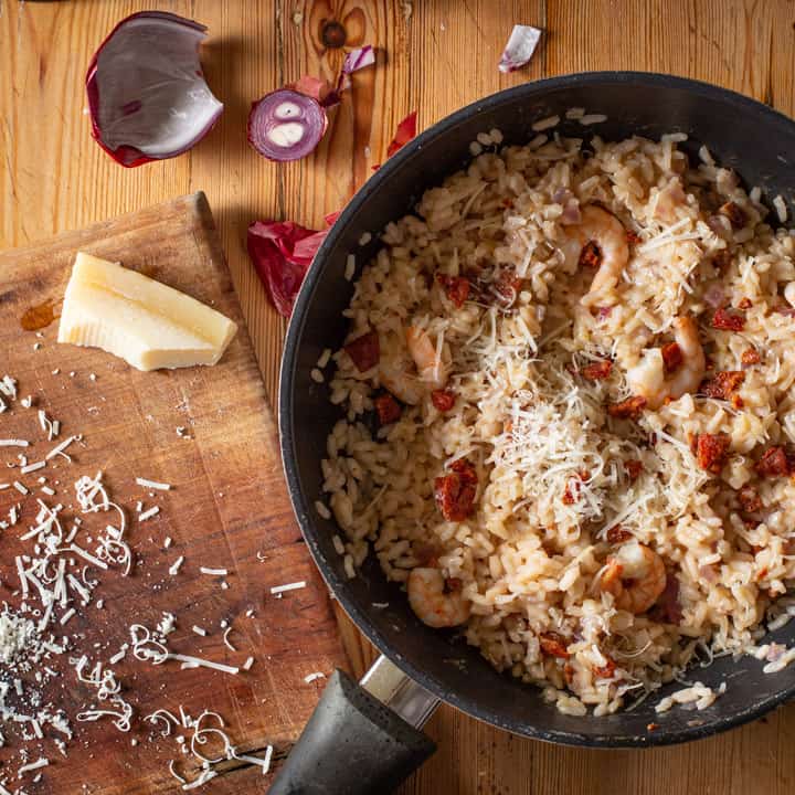 wooden background with black pan of risotto, parmesan rind, messy grated cheese and red onion skin