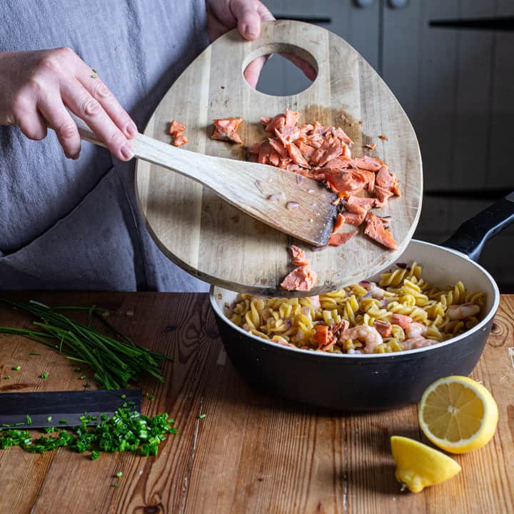 womans hands holding a wooden chopping board of flaked wild salmon over a pan of pasta