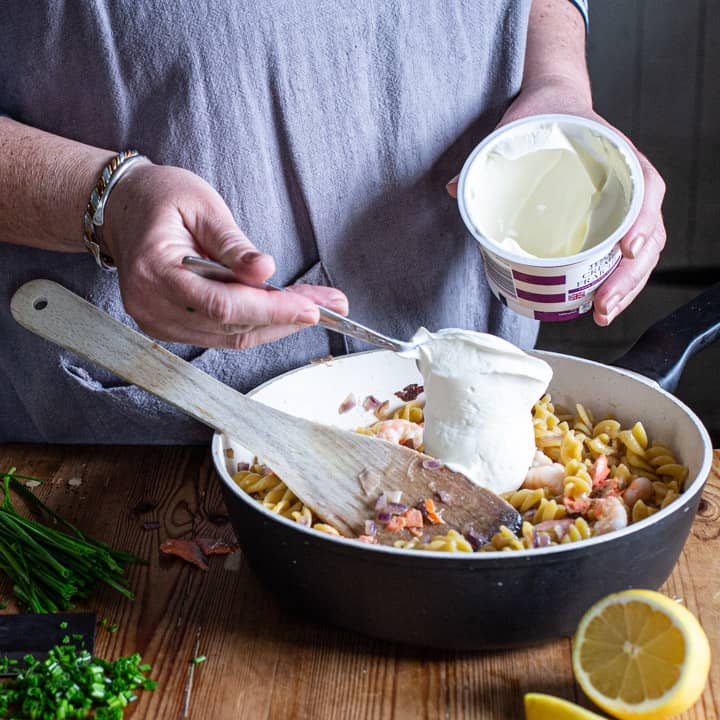 womans hands spooning creme fraiche into a pan of cooked pasta