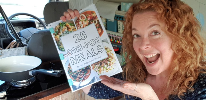 woman in VW campervan kitchen holding a printed A4 ebook called 25 one-pot meals