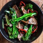 wooden background with black bowl of stir fried broccolini, strips of juicy seared sirloin steak and slices of red chilli