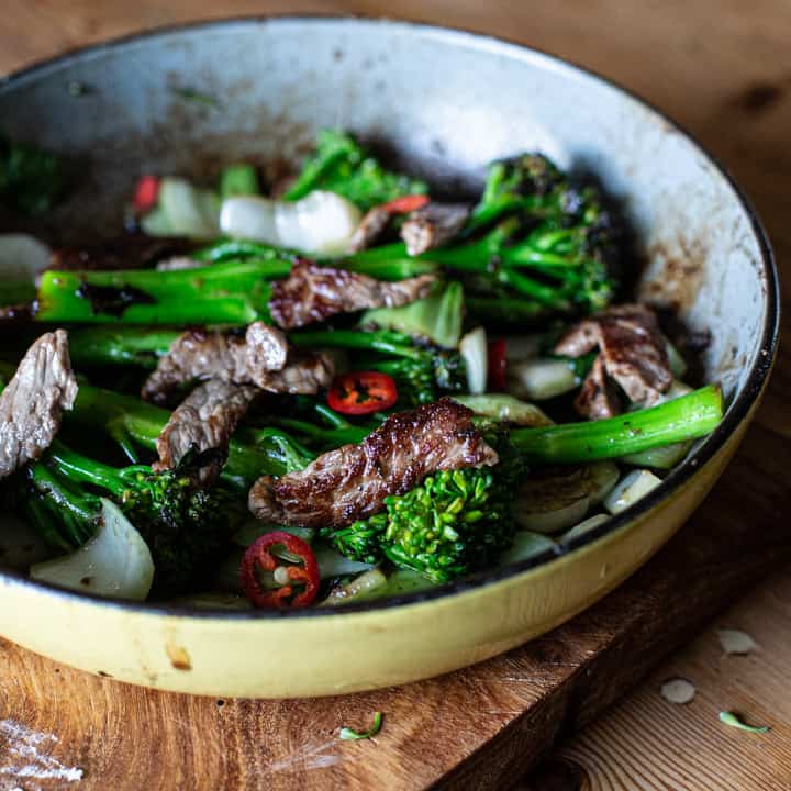 yellow enamel frying pan on wooden surface holding bright green broccolini charred sirloin steak strips and rings of red chilli