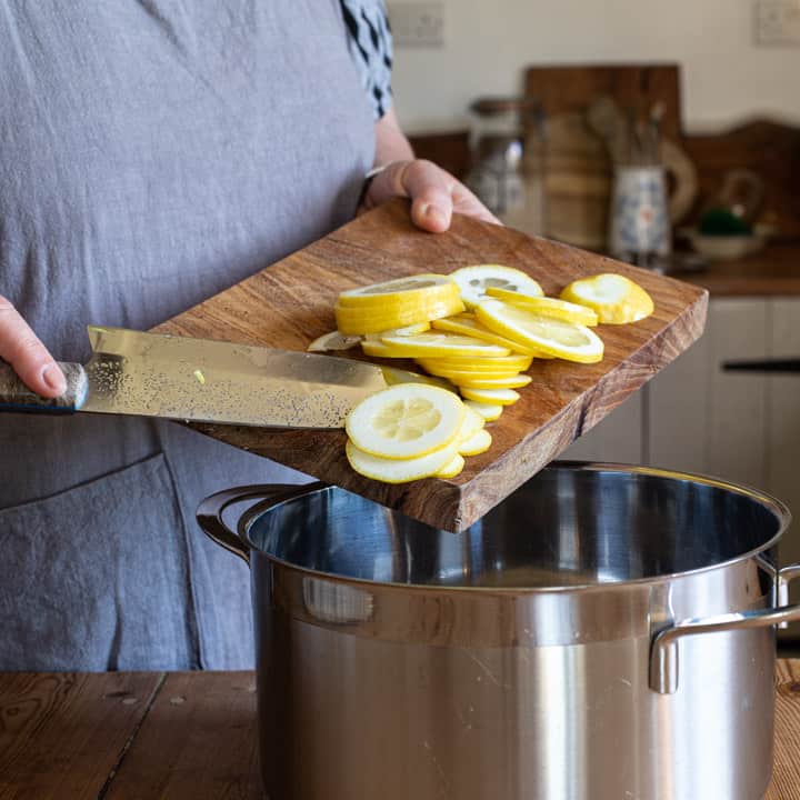 woman in grey sliding several slices of fresh lemon from a wooden board into a large silver saucepan