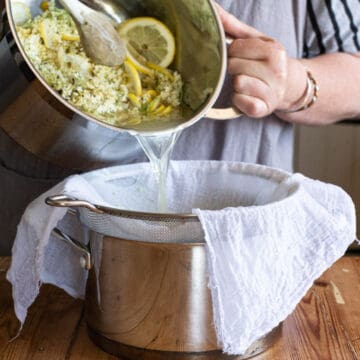 woman tipping liquid, white elderflowers and lemon slices from a large saucepan into a sieve lined with white muslin cloth