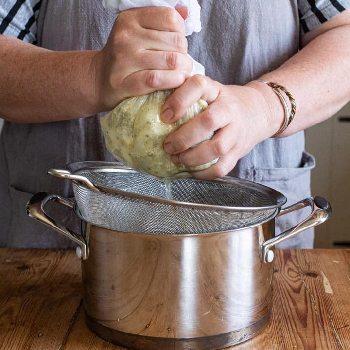 womans hands squeezing the juice from a piece of muslin cloth filled with elderflowers and lemons into a sieve over a silver saucepan