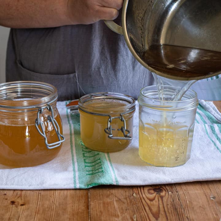 woman in grey pouring liquid jam from a silver saucepan into 3 glass jars on a white cloth