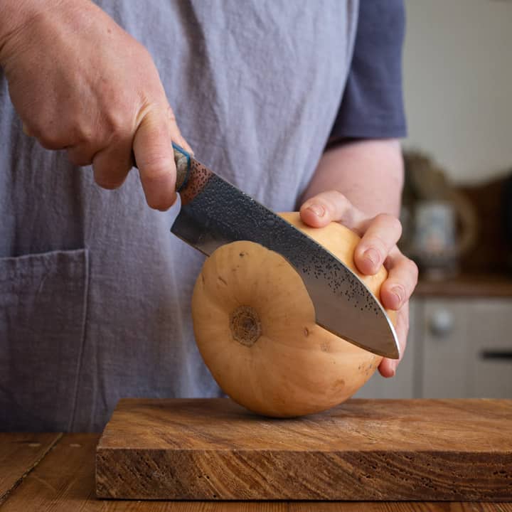 woman in grey cutting a butternut squash with a large knife on a wooden cutting board