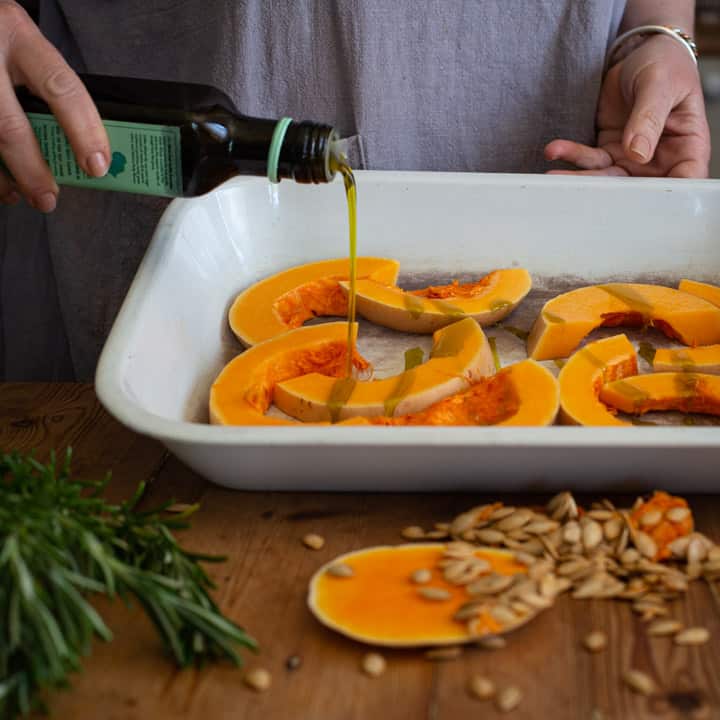 womans hands pouring avocado oil over slices of butternut squash in a white roasting tin